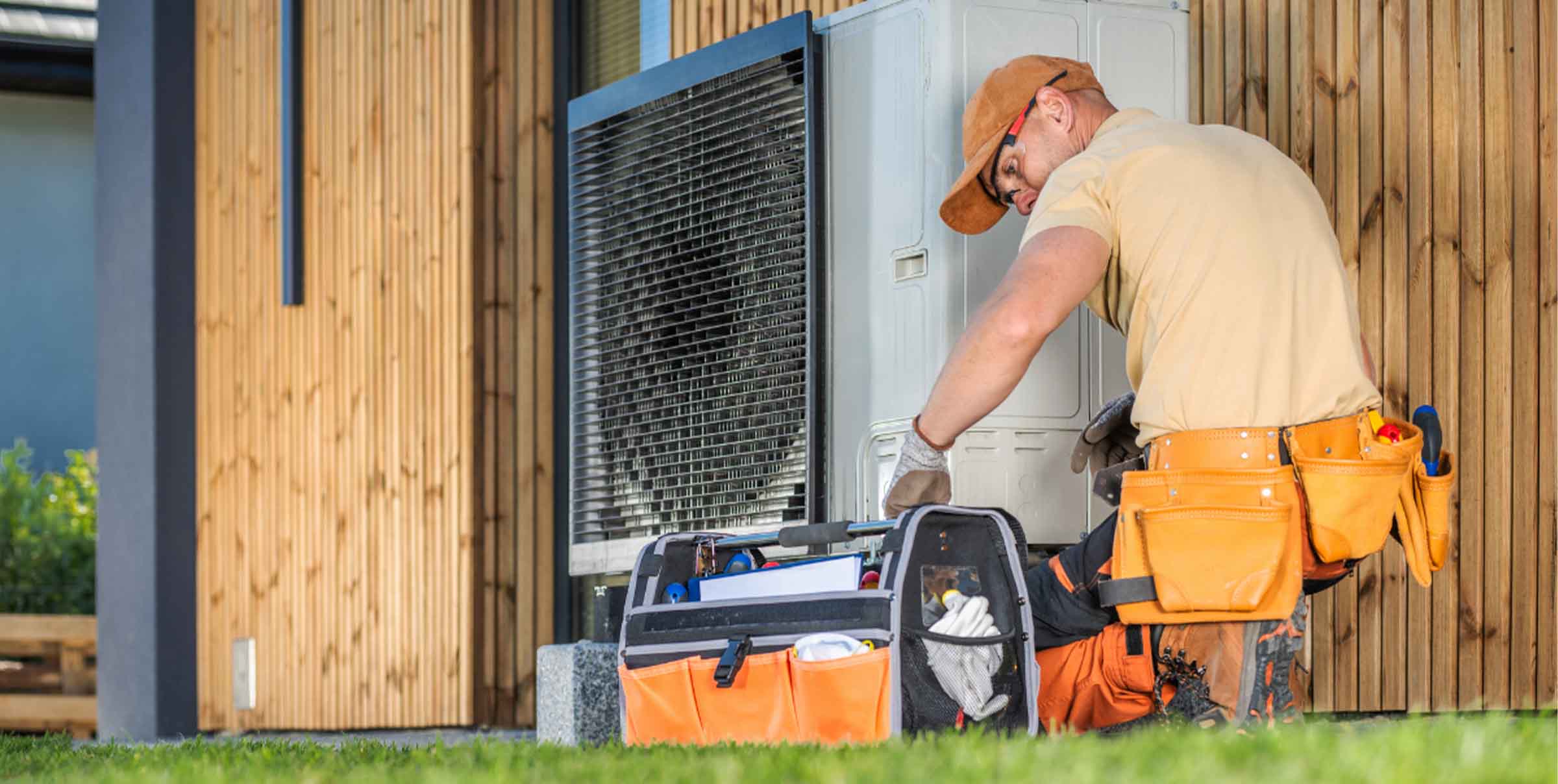 HVAC technician in a tan shirt and tool belt repairing an outdoor heat pump unit against a modern wood-paneled house.